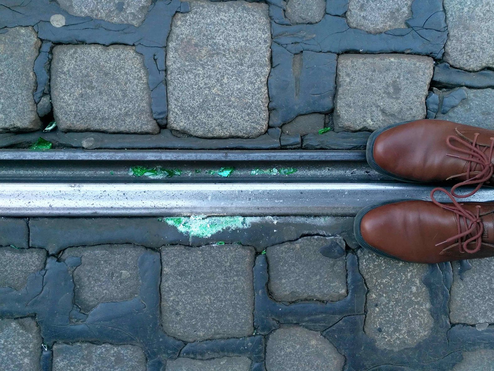 Grüne Glasspillter in und neben einer Straßenbahnschiene, die durch Kopfsteinpflasterung führt. Fotografiert von oben, die Schuhe des Fotografen sind erkennbar.